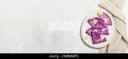Dünne lavendelfarbene Pfannkuchen mit Bananenscheiben und süßem Belag. Gesundes veganes Lactose- und glutenfreies Essen, Frühstückskonzept. Trendiges, hartes Licht, Dar Stockfoto