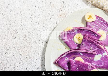 Dünne lavendelfarbene Pfannkuchen mit Bananenscheiben und süßem Belag. Gesundes veganes Lactose- und glutenfreies Essen, Frühstückskonzept. Trendiges, hartes Licht, Dar Stockfoto