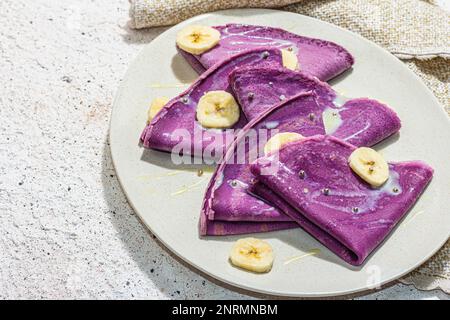 Dünne lavendelfarbene Pfannkuchen mit Bananenscheiben und süßem Belag. Gesundes veganes Lactose- und glutenfreies Essen, Frühstückskonzept. Trendiges, hartes Licht, Dar Stockfoto