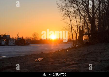 Der Müll taute im Frühling vom Schnee auf dem Eis des Flusses bei Sonnenuntergang auf der Straße in der Stadt Russland auf Stockfoto