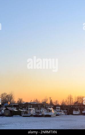 Die strahlend orangefarbene Sonne scheint auf dem Winterparkplatz der Schiffe im Frühling bei Sonnenuntergang in Russland. Stockfoto