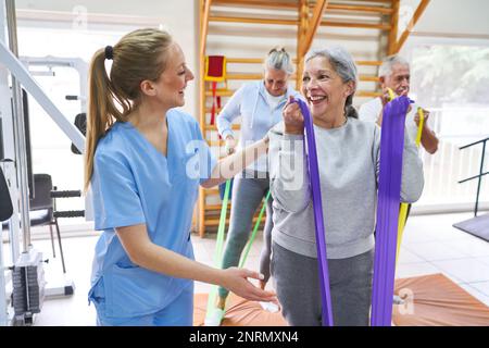 Lächelnde junge Therapeutin, die Seniorin beim Training mit dem Resistenzband im Pflegeheim unterstützt Stockfoto