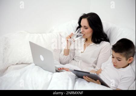 Diät Gewichtsverlust Frau trinkt Wasser im Bett während der Arbeit auf Computer Sohn spielt auf Tablet richtige Ernährung gesunde Lebensweise krank zu Hause Coronavirus Quarantäne Ruhe nach der Arbeit verbringen Zeit Stockfoto