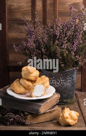 Still Life in der Provence im Herbst mit einer Gruppe französischer Desserts, weißer Eclairs auf dem Teller, Heideblumen und alten Büchern. Braun, Holz, rustikal R Stockfoto