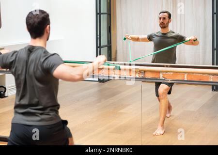 Rückansicht des konzentrierten jungen bärtigen männlichen Athleten in Sportswear, der das Widerstandsband zieht, während er vor dem Spiegel die Schulterstabilität trainiert Stockfoto
