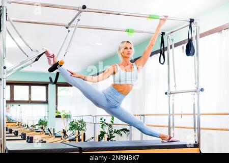 Der ganze Körper einer erwachsenen Sportlerin in blauer Sportbekleidung, die auf dem Pilates Cadillac im Fitnessstudio splittert und wegschaut Stockfoto