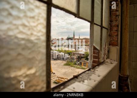 Verlassenes Industriegebäude: Zerbrochenes Fachwerkfenster, durch das ein Industriegelände und die Türme von Lübeck zu sehen sind. Stockfoto