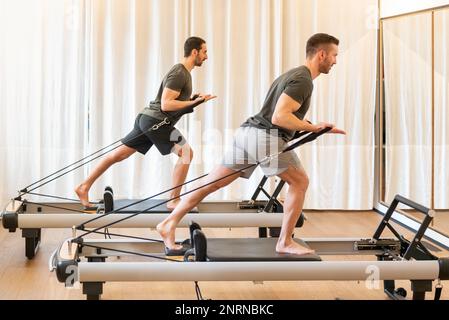 Ganzkörper-Seitenansicht von konzentrierten männlichen Sportlern in aktiver Kleidung, die Stehbaumumarmungen Pilates Training auf professionellen Geräten im Fitnessstudio Stockfoto