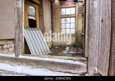 Verlassenes Wohnhaus: Heller Eingangsbereich mit zwei verfallenen Türen, fotografiert durch ein offenes Holzfenster. Stockfoto