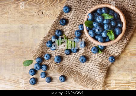 Frisch gepflückte, saftige und frische Blaubeeren mit grünen Minzblättern in einer hellen Holzschüssel auf einem altmodischen rustikalen Tisch aus Jutetuch. Heidelbeere in der Horizontalen Stockfoto