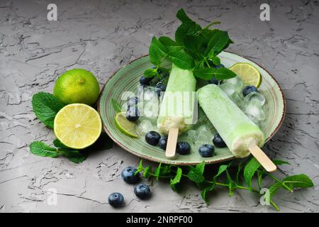 Hausgemachte Kiwi, Limette, Minzeis oder Eis am Stiel mit reifen Blaubeeren auf grünem Teller und grauem Betontisch. Gefrorener Fruchtsaft im Vintage-Stil, clos Stockfoto