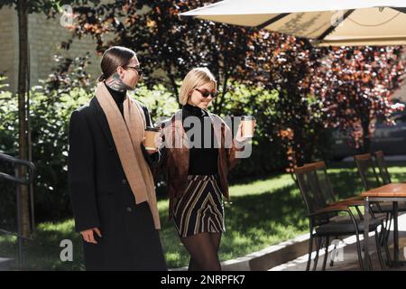 Ein glückliches Paar in einer trendigen Sonnenbrille, das Papierbecher hielt, während es draußen spazierte, ein Stockbild Stockfoto