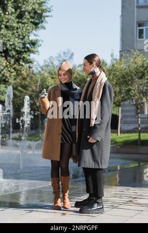 Die ganze Länge einer glücklichen blonden Frau im Mantel und Hut steht mit tätowiertem Freund draußen, Bild der Aktie Stockfoto