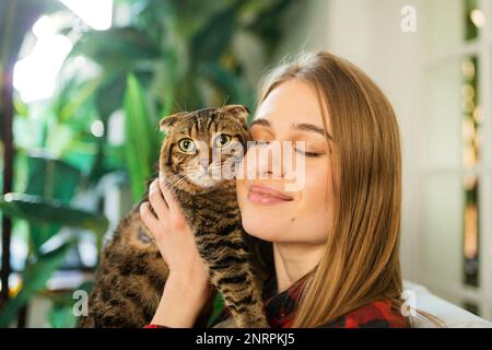 Lässige junge blonde Frau hält Schulter eine süße, grün-äugige schottische Tabby-Katze und umarmt sie zu Hause. Das Konzept der Liebe und Fürsorge für Haustiere Stockfoto