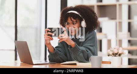 Aufgeregte junge schwarze afrikanische Frau trägt Kopfhörer und genießt Spiele über die App auf dem Mobiltelefon. Stockfoto