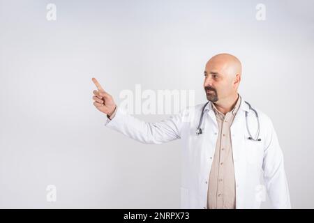 Lächelnder Arzt zeigt Platz zum Kopieren. Weißer, gutaussehender, männlicher Arzt in weißem Mantel mit Stethoskop, das mit seinem Finger auf einen leeren Raum zeigt. Isoliertes Grau. Stockfoto