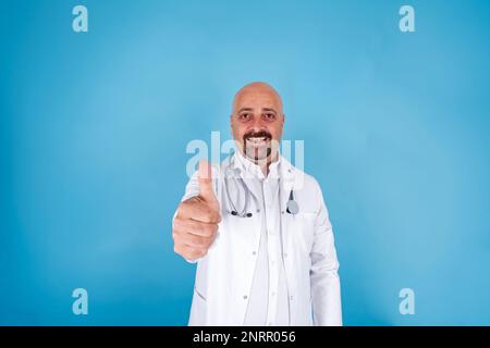 Lächelnder männlicher Arzt. Er steht auf blauem Hintergrund. Er trägt einen weißen Mantel mit Stethoskop und zeigt Daumen nach oben. Gesundheitswesen, medizinisches Konzept. Stockfoto