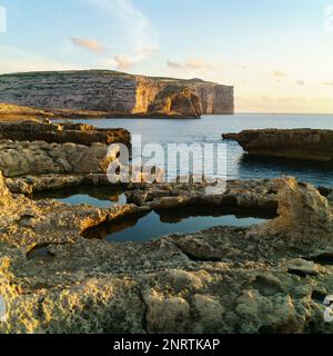 Dwejra, Insel Gozo, Malta. Atemberaubender Sonnenaufgang in der Nähe der Azure Window Ruinen. Warmes gelbes Sonnenlicht auf den Felsen und dem Meer, blau mit Wolken. Beruhige mich Stockfoto