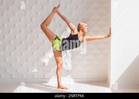 Fit hübsche junge Frau mit schwarzem Oberteil und grünen Shorts, die Yoga-Stretching-Übungen im Fitness-Training in einem weißen Fitnessstudio mit Dayling s macht Stockfoto