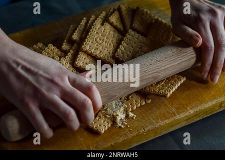 Hände, die Kekse mit einer Walznadel zerbrechen, um die knusprige Basis eines Käsekuchen zuzubereiten Stockfoto