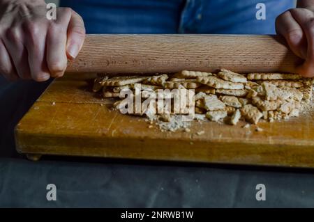 Hände, die Kekse mit einer Walznadel zerbrechen, um die knusprige Basis eines Käsekuchen zuzubereiten Stockfoto