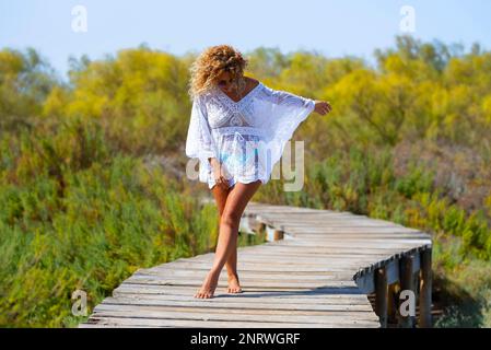 Eine wunderschöne Frau genießt den Sommerurlaub und das Reiseziel, das sich auf einem hölzernen Pier mit weißem, elegantem Strandkleid und blondem Cu ausgibt Stockfoto