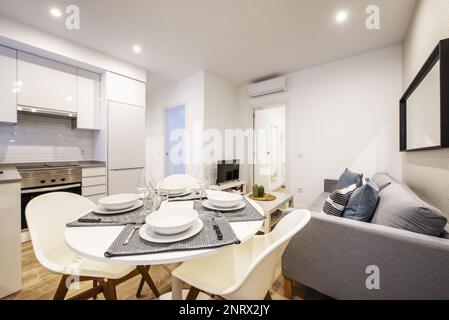 Studio-Apartment mit offener Küche und rundem Esstisch aus weißem Holz mit Service und grauem Sofa für 3 Personen Stockfoto