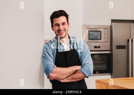 Attraktiver weißer Mann mit einer Schürze, der in der Küche steht, lächelt und Arme kreuzte und in die Kamera schaute. Vorderansicht eines glücklichen, gutaussehenden Mannes, der zu Hause an einer Wand lehnt. Hochwertiges Foto Stockfoto