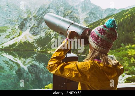 Junge blonde Frau mit gelber Jacke, lila und blauer Mütze, die durch ein Münzfernglas auf Berge und See blickt. Kopierraum, Rückseite V Stockfoto