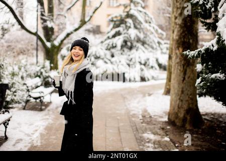 Eine junge Frau steht in einem Winterwunderland, trägt warme Kleidung und trinkt heißen Kaffee zum Mitnehmen Stockfoto