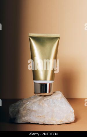 Goldene, cremefarbene Röhre auf natürlichem Podiumständer. Hautpflege, Beauty und Spa-Konzept. Beiger Hintergrund mit Licht und Schatten. Stockfoto