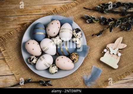 Grauer Teller mit bemalten Ostereiern und Weidenzweigen auf einem Holztisch. Ostergrußkarte. Draufsicht, flach liegend. Stockfoto