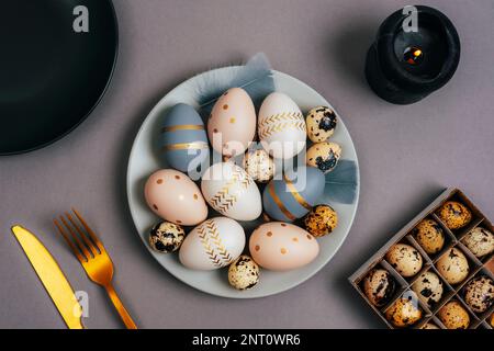 Grauer Teller mit lackierten Ostereiern, Kerze und Gabel und Messer auf grauem Hintergrund. Ostertisch. Draufsicht, flach liegend. Stockfoto