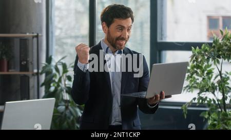 Glücklicher, aufgeregter Geschäftsmann im Büro, der seine E-Mails auf dem Laptop liest gute Nachrichten, Mann Gewinner erfolgreicher Unternehmer, erhält Bonusprämie zum Feiern der Belohnung Stockfoto
