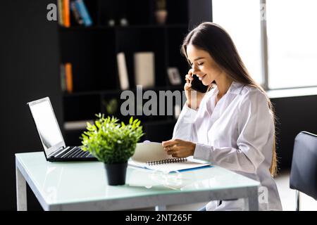 Süße junge Erwachsene mit einem Grinsen am Telefon, während sie am Laptop am Schreibtisch neben der Kaffeetasse mit großem Fenster im Hintergrund arbeiten Stockfoto