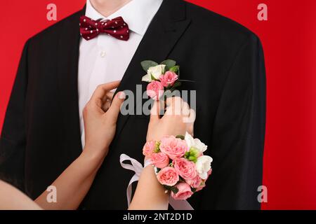 Junge Frau, die Ansteckblume auf dem Anzug ihres Abschlussball-Dates auf rotem Hintergrund anklebt, Nahaufnahme Stockfoto