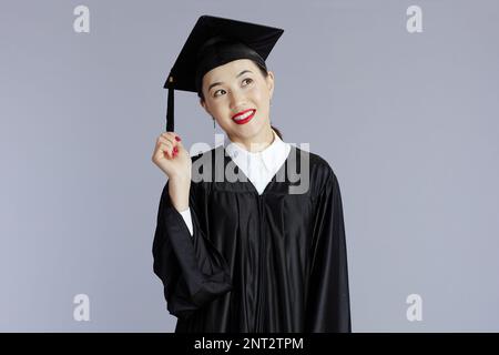 Pensive, moderne asiatische Studentin in Abschlusskleid mit Mütze, die Quasten vor grauem Hintergrund hält. Stockfoto