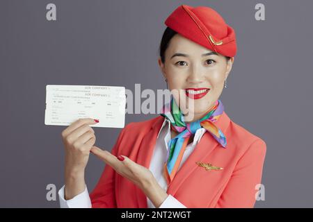 Fröhliche, stilvolle Stewardess Asiatin in rotem Rock, Jacke und Hut Uniform mit Flugtickets isoliert auf grauem Hintergrund. Stockfoto