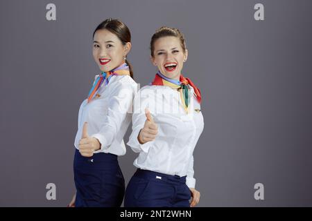 Fröhliche, moderne Flugbegleiterinnen mit blauem Rock, weißem Hemd und Schal, die Daumen nach oben vor grauem Hintergrund zeigen. Stockfoto