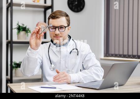 Junger attraktiver Hals-Nasen-Ohren-Arzt, der an seinem Arbeitsplatz in der Praxis sitzt und einen Nasenspiegel in der Hand hält. Stockfoto