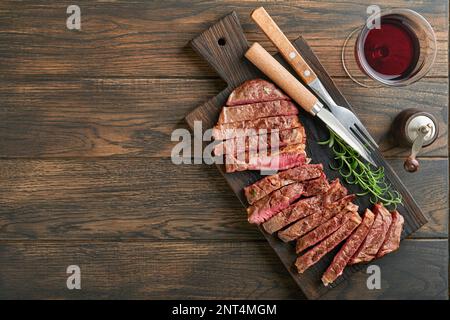 Steaks. Geschnittenes gegrilltes Fleischsteak New York oder Ribeye mit Gewürzen, Rosmarin und Pfeffer auf schwarzem Marmorbrett auf altem Holzhintergrund. Draufsicht. Modell Stockfoto