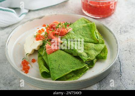 Grüne Pfannkuchen. Runde, dünne, grüne Spinat-Crepe-Pfannkuchen mit rotem und schwarzem Kaviar und Joghurt-Sauce auf grauem Betonhintergrund. Gesundes veganes Frühstück Stockfoto