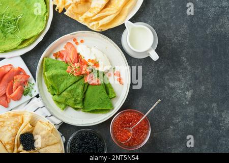 Verschiedene grüne und weiße Pfannkuchen. Dünner grüner Spinat und weiße Crepe-Pfannkuchen mit rotem und schwarzem Kaviar, Sauce auf grauem Beton-Tischbrea Stockfoto