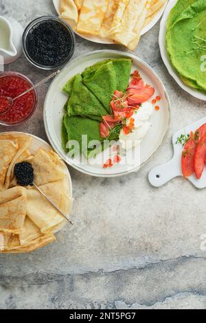 Verschiedene grüne und weiße Pfannkuchen. Dünner grüner Spinat und weiße Crepe-Pfannkuchen mit rotem und schwarzem Kaviar, Sauce auf grauem Beton-Tischbrea Stockfoto