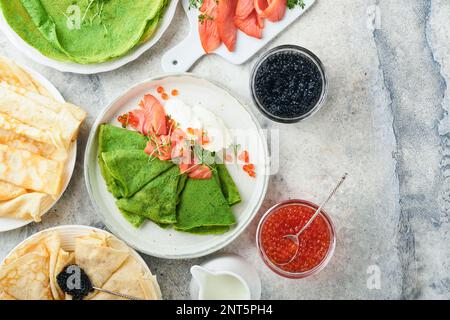 Verschiedene grüne und weiße Pfannkuchen. Dünner grüner Spinat und weiße Crepe-Pfannkuchen mit rotem und schwarzem Kaviar, Sauce auf grauem Beton-Tischbrea Stockfoto