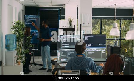 Ein vielseitiges Team von Software-Ingenieuren, die Code auf einem Wandbildschirm analysieren, mit einem digitalen Tablet arbeiten und mit künstlicher Intelligenz arbeiten. Programmierer analysieren Cloud-Computing-Projekte mit Big Data. Stockfoto
