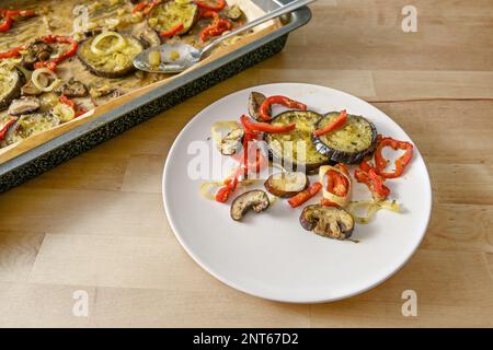 Mediterranes Ofengemüse, in Olivenöl gebacken, mit Kräutern und Gewürzen auf einem Teller und auf einem Backblech, Holztisch, vegetarisches Gericht, Kopierraum, Sel Stockfoto