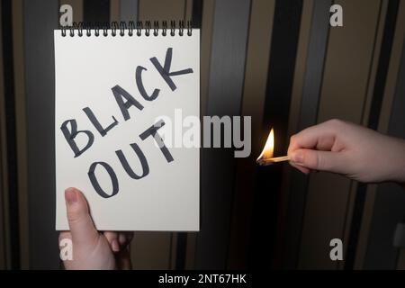 Notizblock mit der Aufschrift „Blackout“ in einer Hand und brennenden Streichhölzern in der anderen Hand (Nahaufnahme). Stromausfallkonzept. Energiekrise Stockfoto