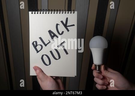 Ein Notizblock mit der Aufschrift „Blackout“ in der einen Hand und eine elektrische Glühbirne in der anderen Hand (Nahaufnahme). Stromausfallkonzept. Energiekrise Stockfoto