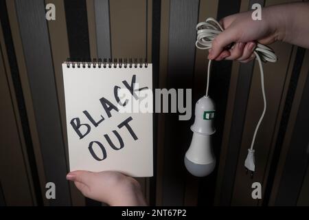 Ein Notizblock mit der Aufschrift „Blackout“ in einer Hand und eine elektrische Glühlampe mit einer Halterung und einem elektrischen Stecker in der anderen Hand (Nahaufnahme) Stockfoto